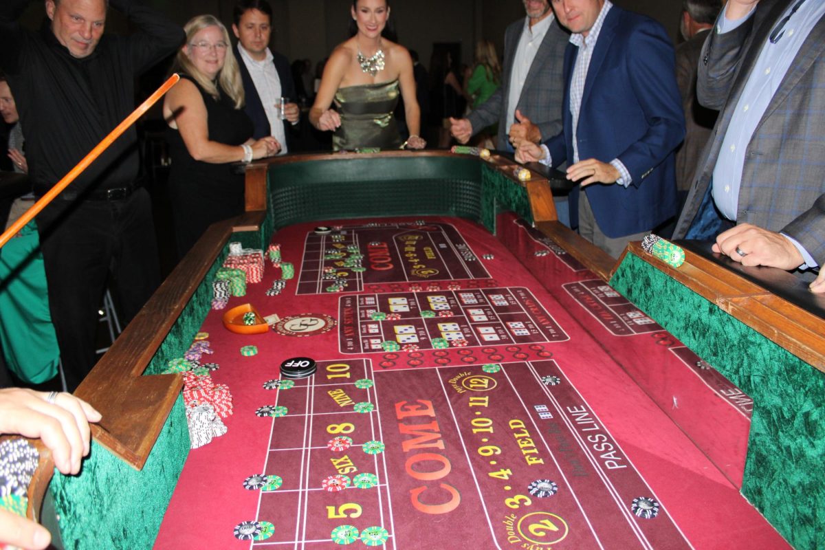 A crowd gathers around the craps table inside the ballroom "casino" anticipating the result of the dice roll.