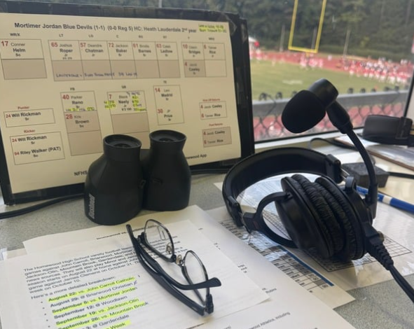 Binoculars, glasses and a headset rest on a table full of research for the home game against Mortimer Jordan. Walker, Brasher and Bloom work to compile statistics on the opposing team to fully flesh out their commentary for viewers at home. 
