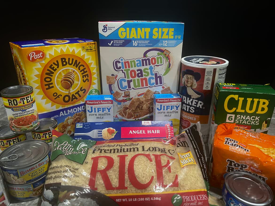 An array of shelf-stable food items sits on a dining room table. Highest priority items for donation include dry goods like rice and cereal, but all non-perishable items are welcome.